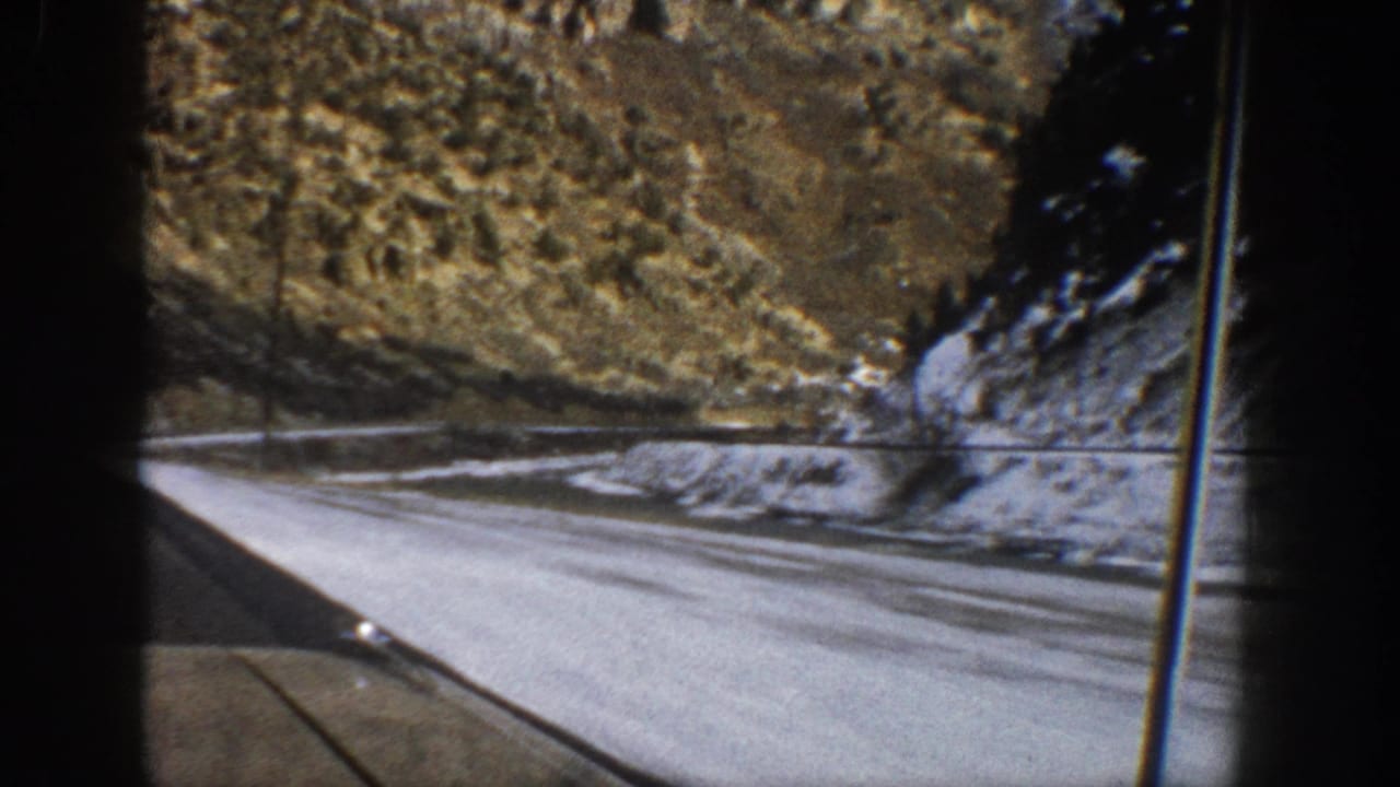 Winding road curves beside snowy ground and a rocky hillside, viewed through a windshield.