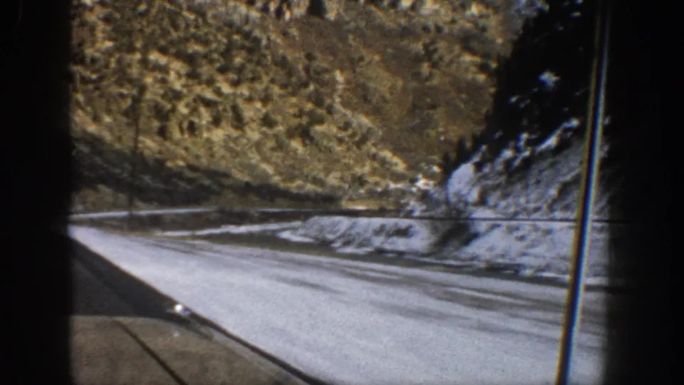 A 1961 Aspen, Colorado road curves through snow-dusted terrain, filmed from a moving vehicle with the windshield edge visible.