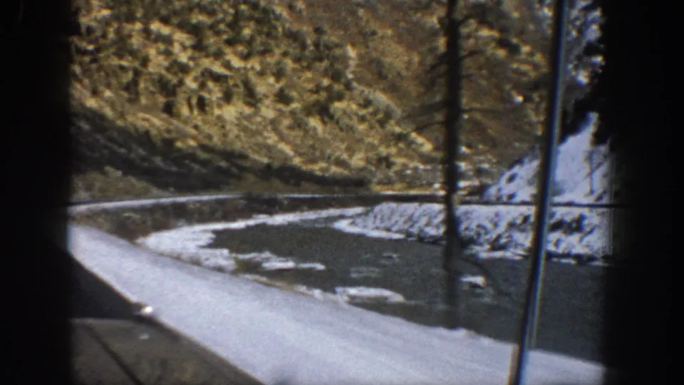 A windshield-framed 1961 Aspen mountain-road view shows a snowy curve, dark roadside shadows, and hills beyond.