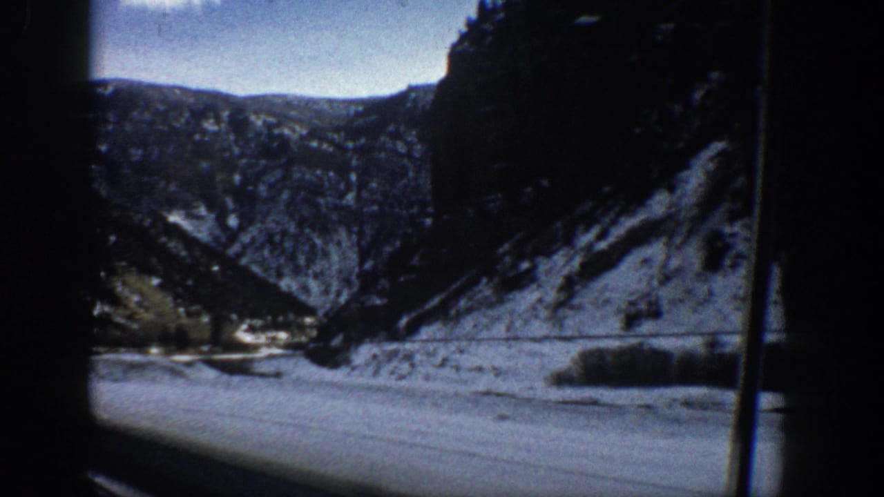 Snow-dusted mountain valley with river and road edge framed by a vertical car-window post.