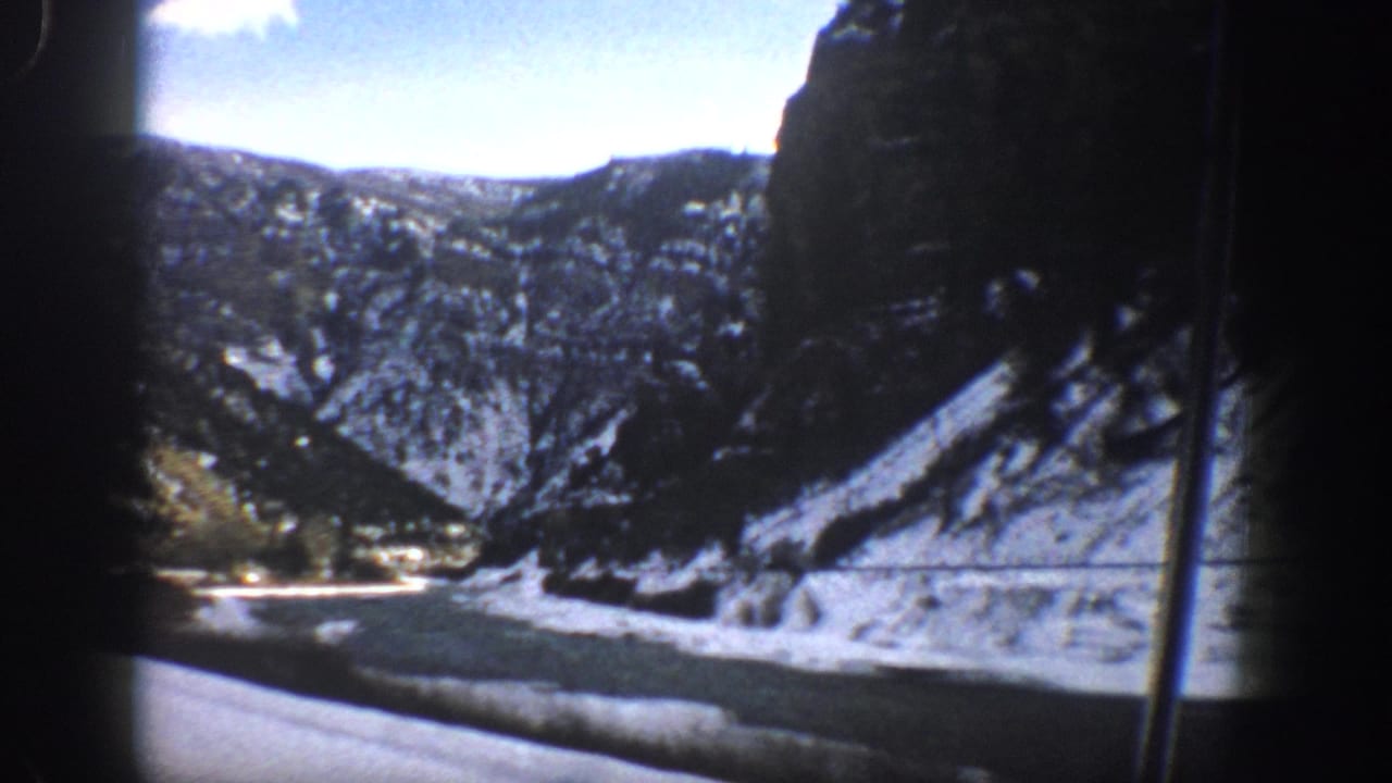 1961 Aspen-area 8mm frame showing a snow-dusted mountain canyon from a moving car, with cliffs, trees, and roadside water visible.