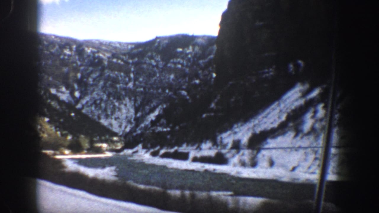 Mountain canyon with snow patches, rocky slopes, trees, and a roadside watercourse seen through film borders.