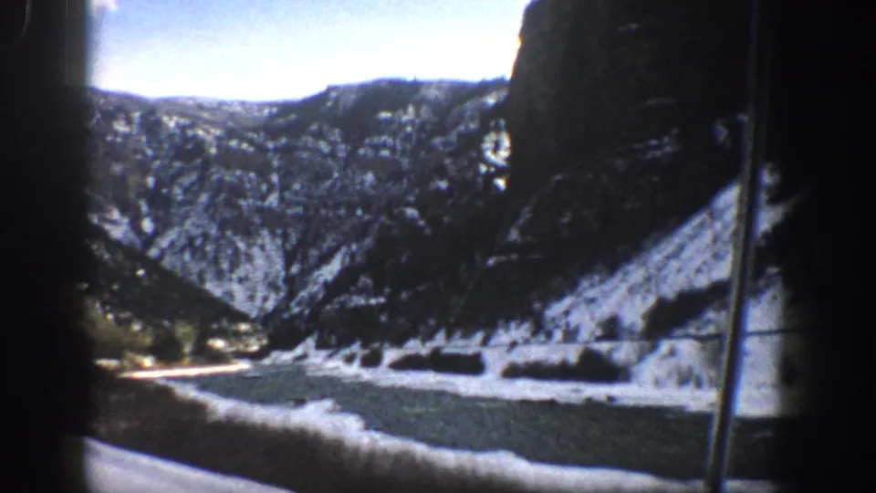 1961 Aspen-area home-movie frame from a car, showing snowy slopes, a canyon wall, trees, and water beside the road.