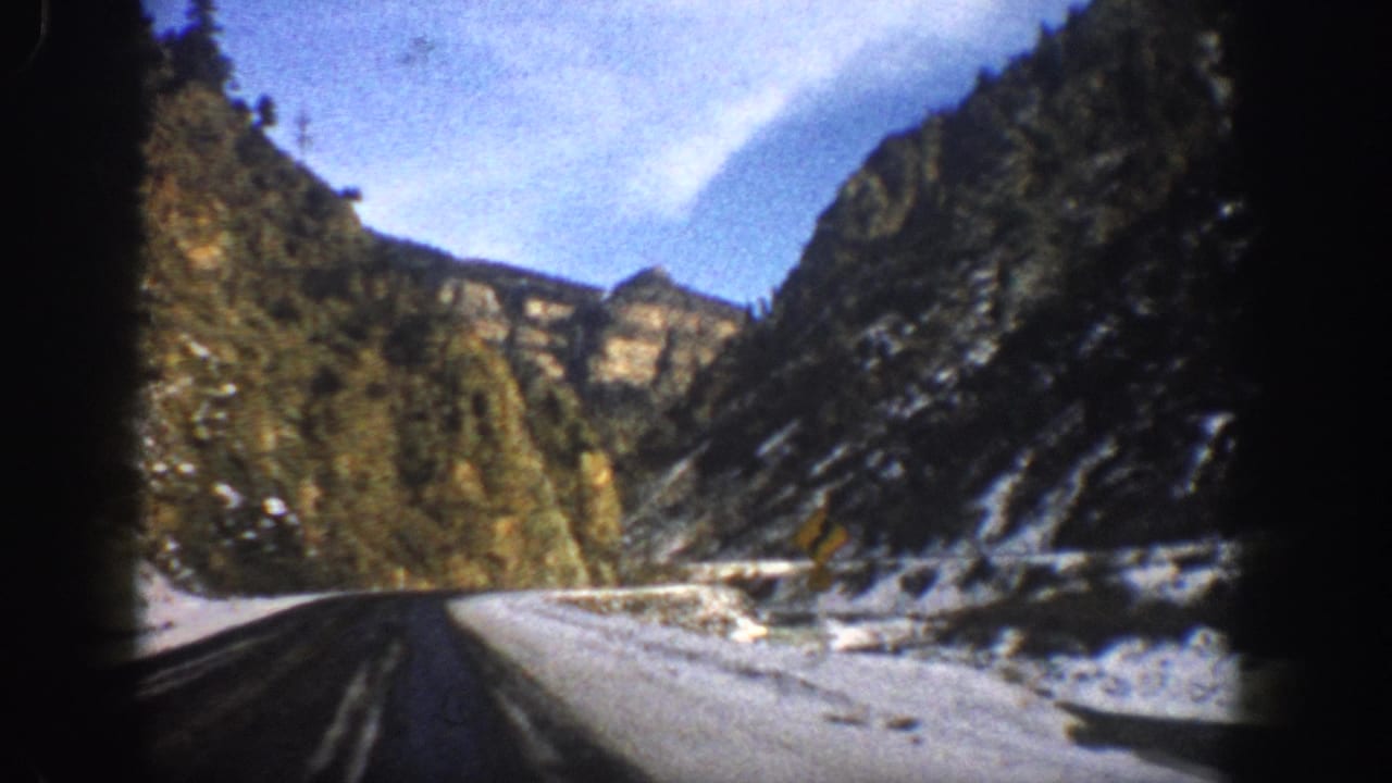 1961 Aspen, Colorado 8mm frame showing a vehicle-eye view along a curving snowy mountain highway between rocky, tree-covered slopes.