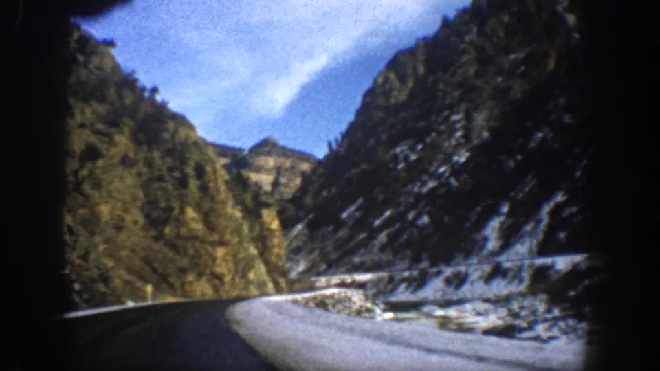 1961 Aspen, Colorado winter driving frame with a curving roadway, snow along the shoulder, rugged canyon walls, and 8mm film-edge darkness.
