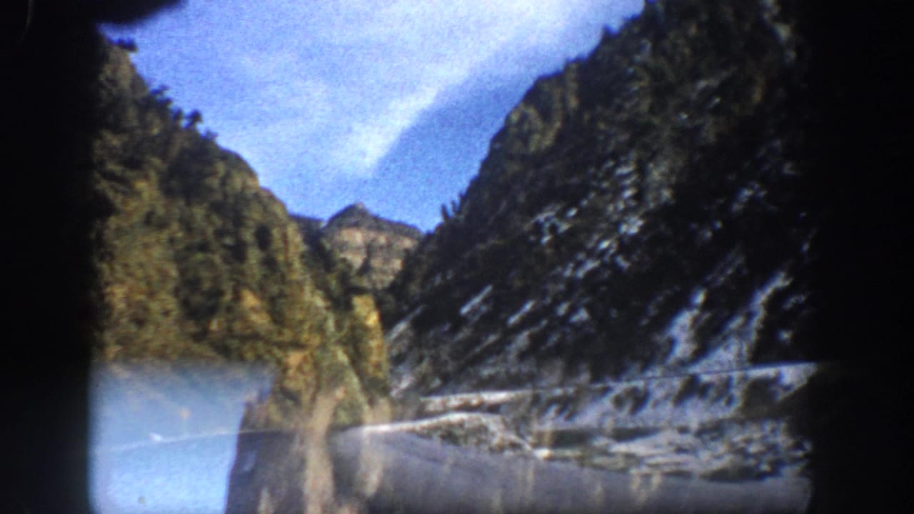 1961 Aspen-area 8mm view from a moving vehicle, showing a mountain road below steep rocky cliffs with snow patches under blue sky.