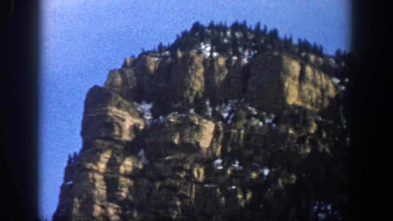 1961 Colorado mountain view with a tall rock face, scattered evergreens, and remaining snow seen through soft 8mm color grain.