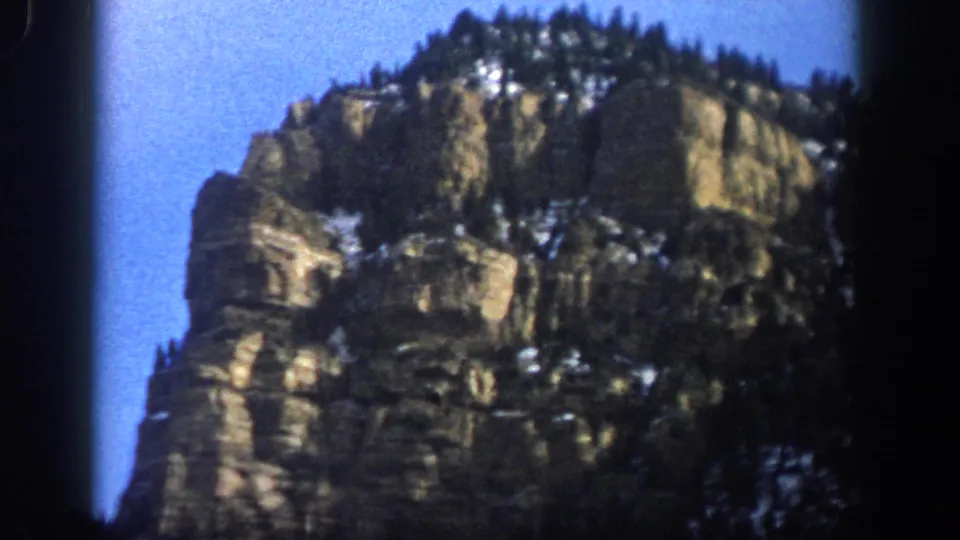 8mm home-movie frame from the Aspen, Colorado clip, showing a rugged cliff face with evergreens and patchy snow in saturated color.