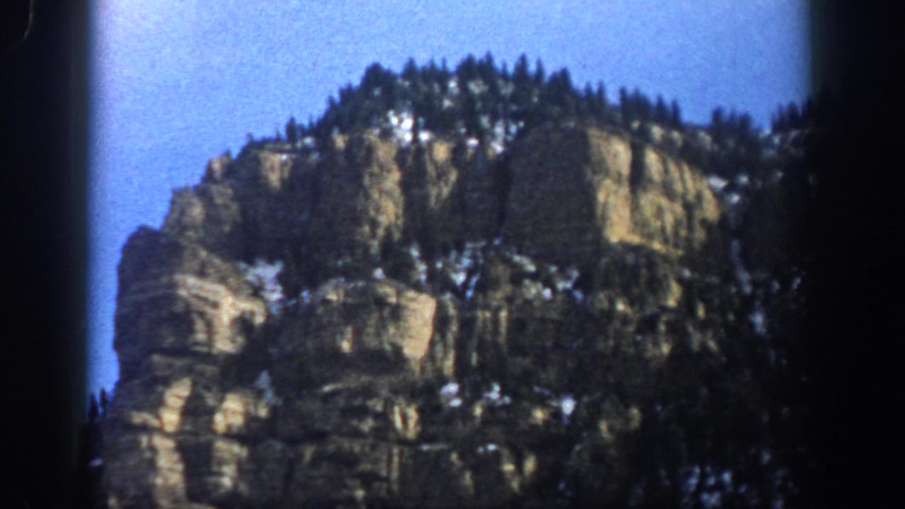 1961 Aspen, Colorado home-movie frame showing rugged cliffs, evergreen forest, snow patches, blue sky, and dark 8mm borders.