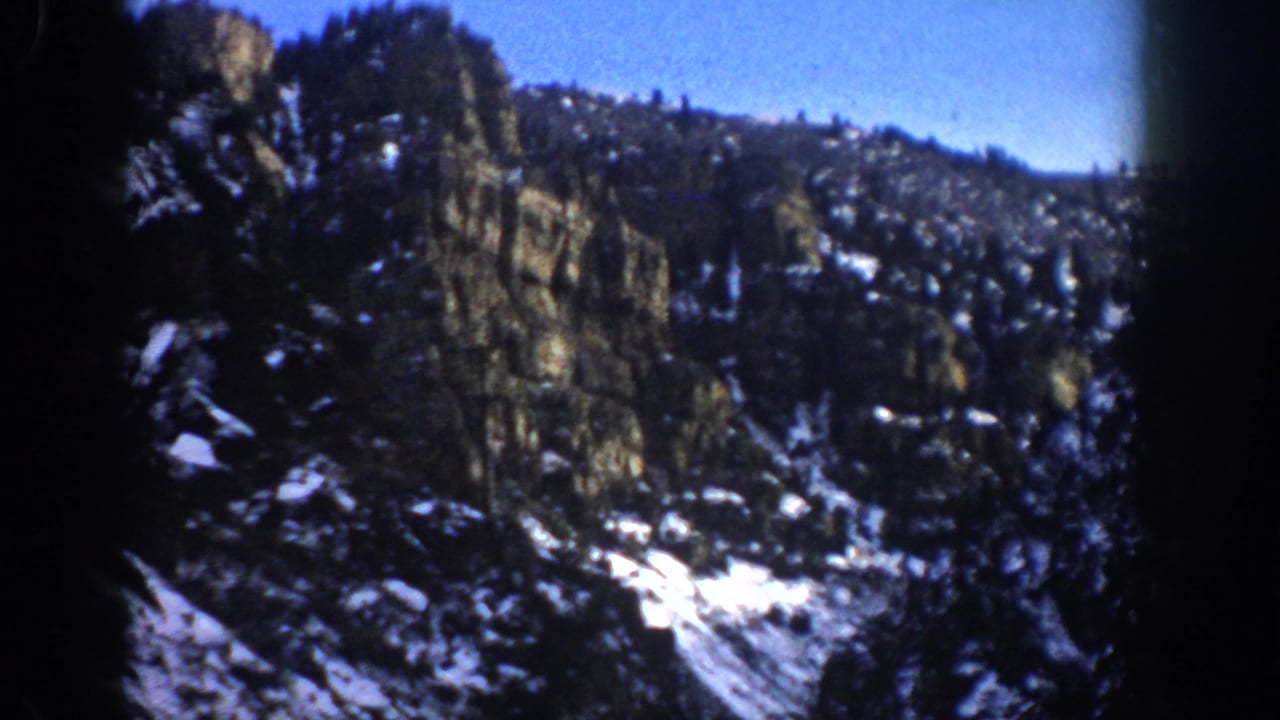 1961 Aspen, Colorado frame with a darker rocky mountainside, evergreen forest, scattered snow, blue sky, and handheld 8mm framing.