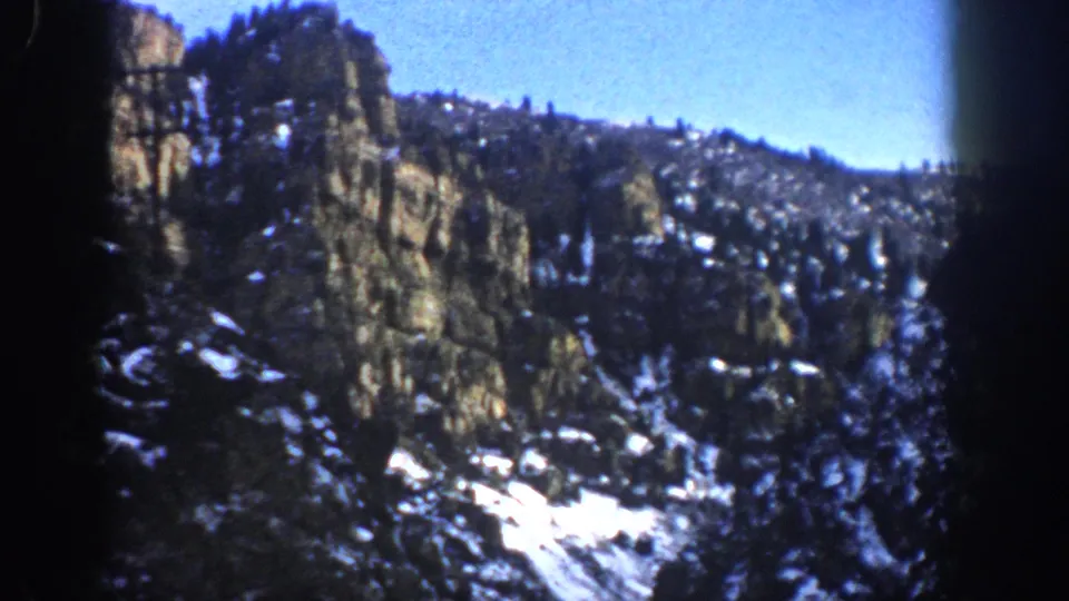 1961 Aspen, Colorado closing frame showing a rugged cliff, dark evergreens, scattered snow, blue sky, and rounded 8mm film edges.