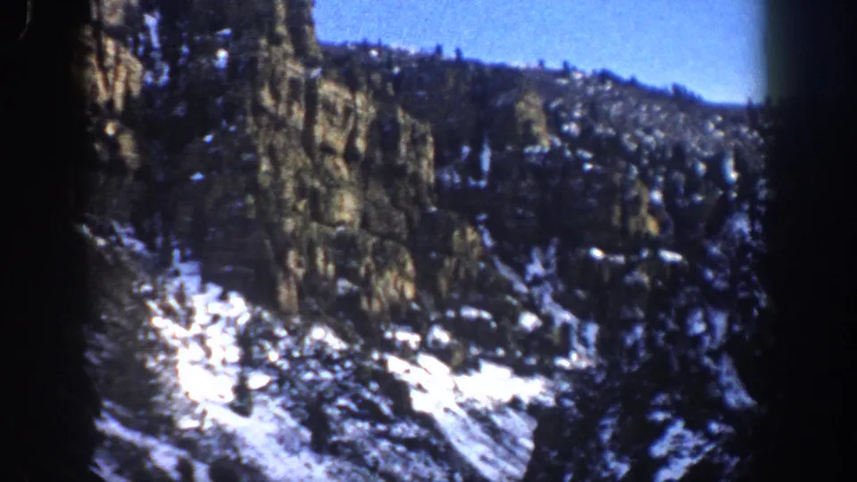 1961 8mm frame from Aspen, Colorado showing a snow-streaked rocky slope, dense evergreens, and visible dark film borders.