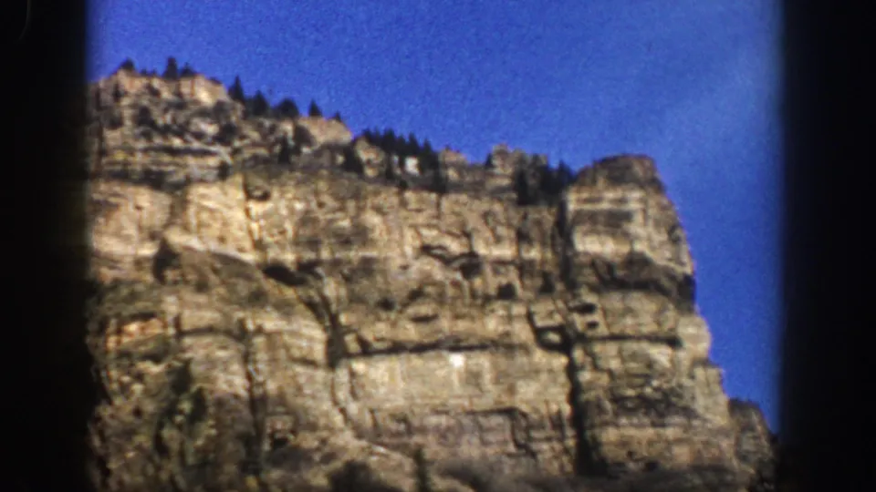 Aspen, Colorado 1961 home-movie frame showing a sunlit cliff formation with trees along the ridge and blue sky behind it.