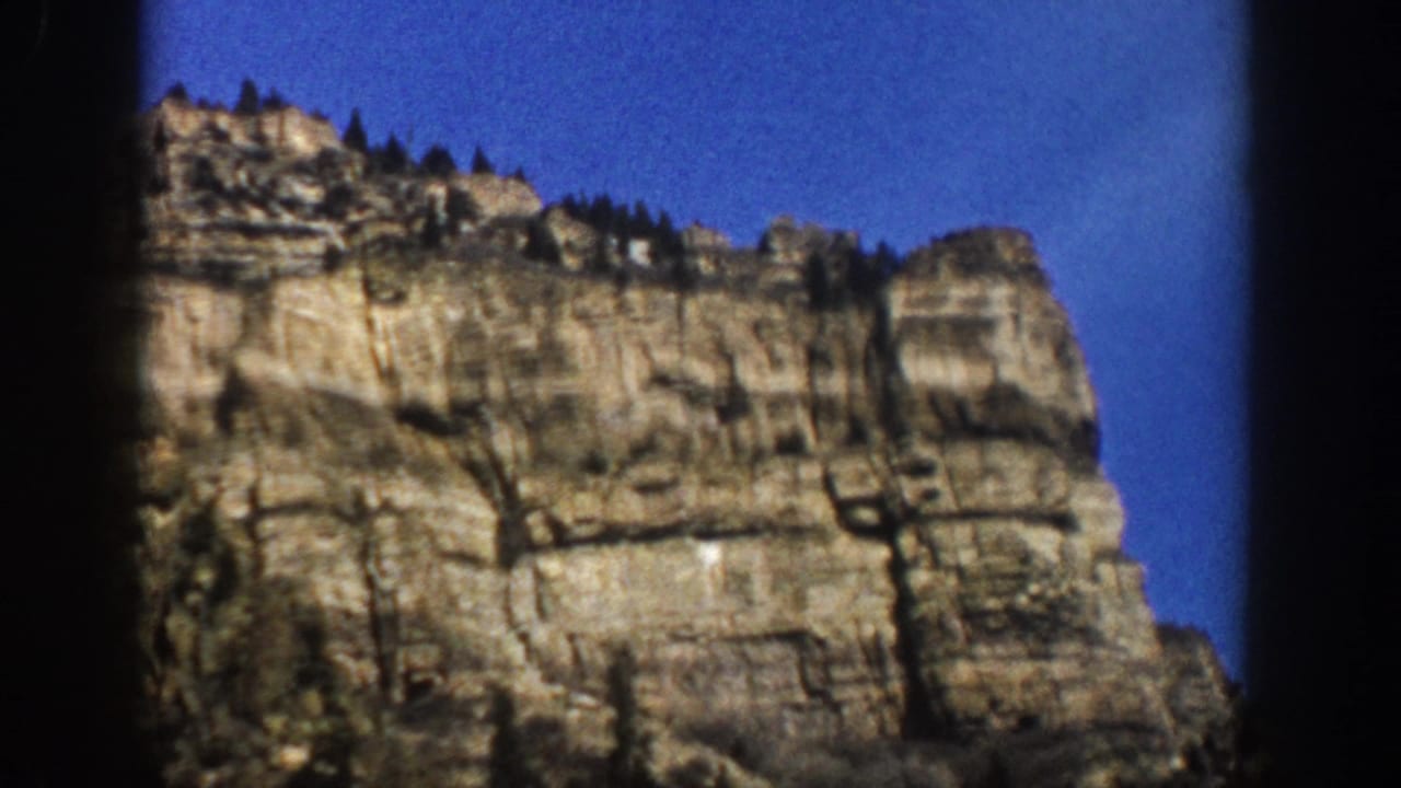 1961 Aspen, Colorado home-movie frame showing a layered rocky cliff beneath a clear blue sky with sparse trees on the ridge.