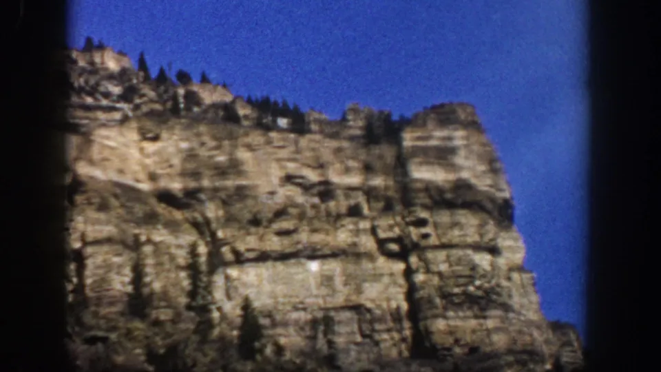1961 Aspen, Colorado home-movie frame showing the cliff face shifted lower in view, with stone layers and trees near the ridge.