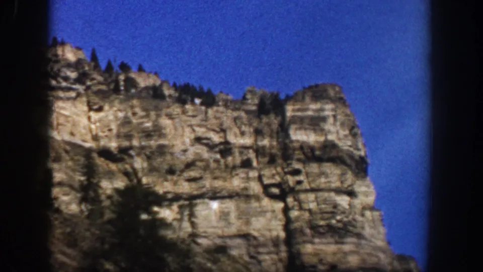 1961 Aspen, Colorado frame showing a large layered cliff, ridgeline evergreens, and strong blue sky within dark 8mm borders.
