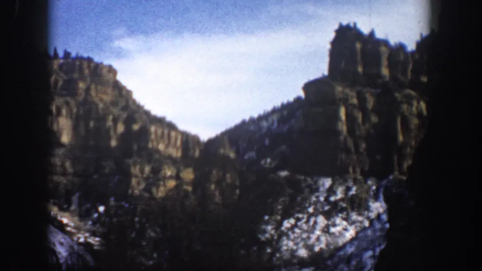 1961 Aspen, Colorado 8mm frame with steep rock towers, dark evergreen slopes, snow patches, and a bright sky in a mountain canyon.