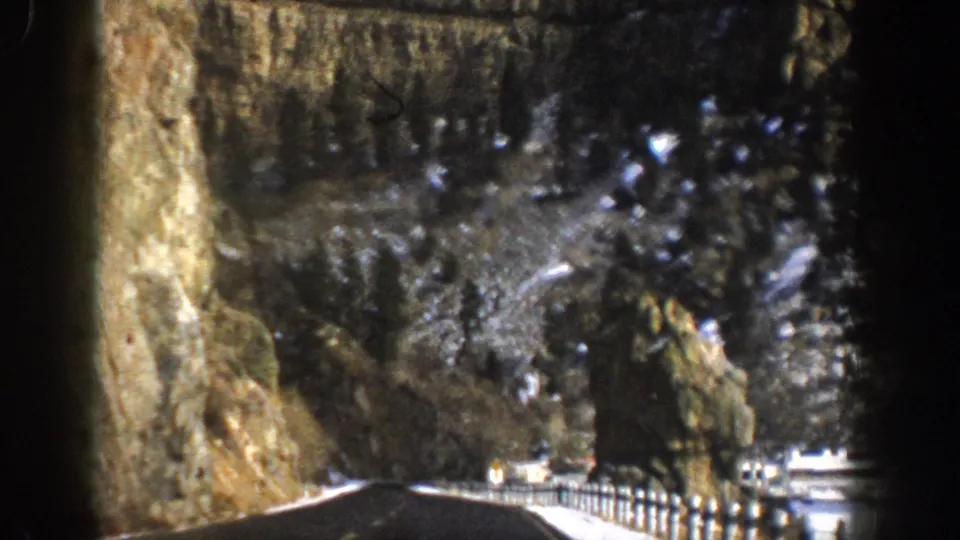 1961 Aspen, Colorado 8mm frame looking down a canyon road past guardrails, a rock spire, snow-dusted slopes, and small distant structures.