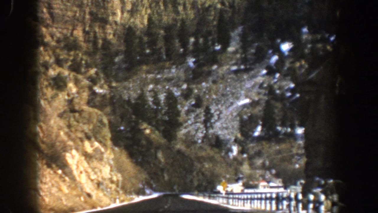 Winding mountain highway with guardrail, cliff face, conifers, snow patches, and a small vehicle.