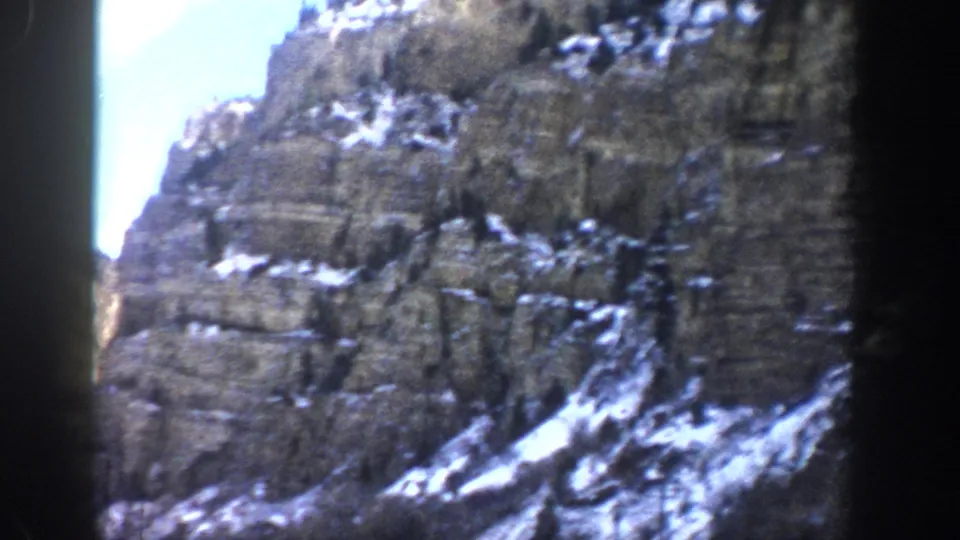 1961 Aspen, Colorado home-movie frame with steep canyon rock walls, scattered snow, and a narrow blue-sky opening.