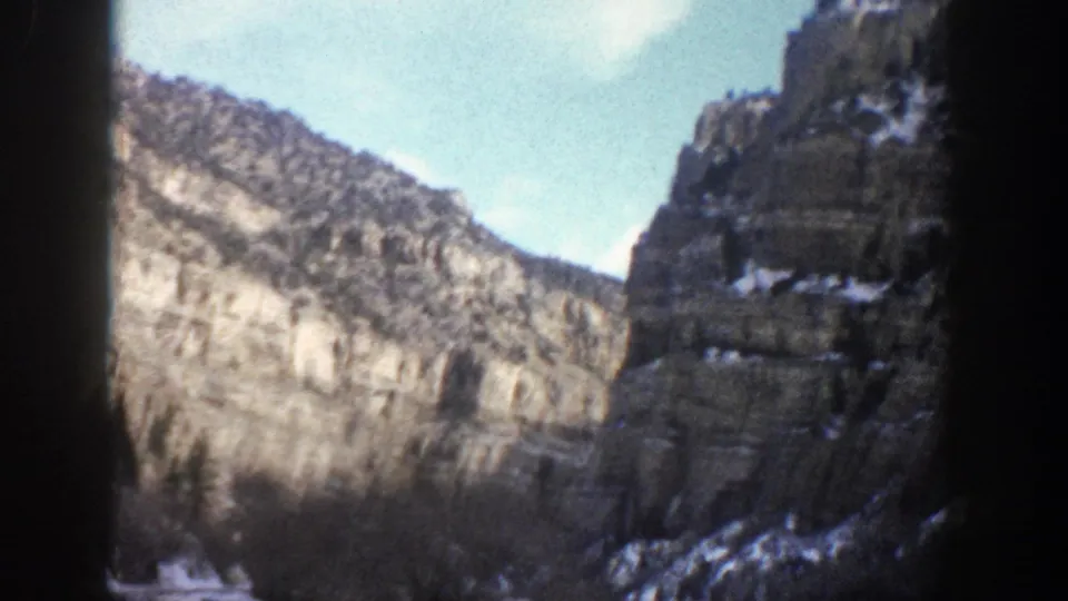 1961 Aspen, Colorado 8mm frame showing broad cliff faces, snow near the lower slopes, evergreen silhouettes, and projector border shadows.