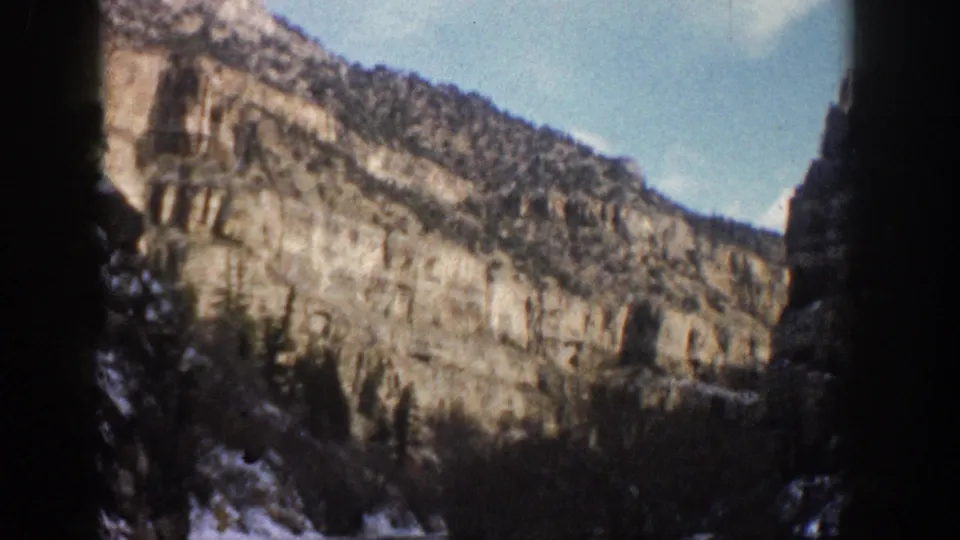 1961 Aspen, Colorado home-movie frame of a sunlit cliff ridge, scattered snow, dark evergreens, and blue sky within 8mm frame edges.