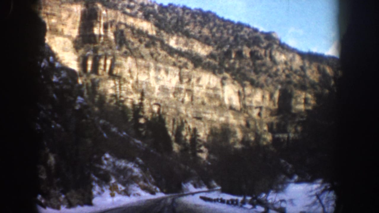 Aspen, Colorado home-movie frame showing a snowy road corridor beneath pale stratified cliffs and scattered evergreens.