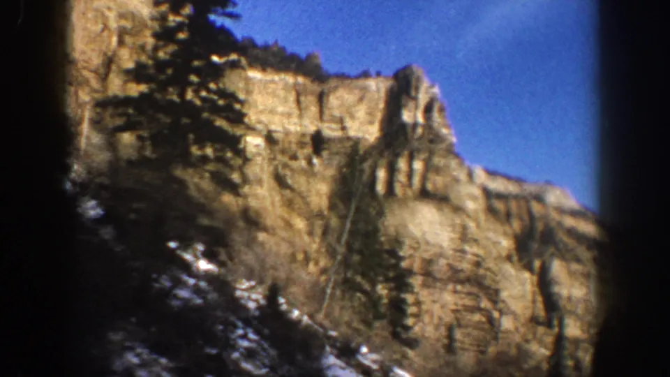 1961 8mm travel footage frame with a close cliff face, evergreens, snow patches, and dark car-window framing.