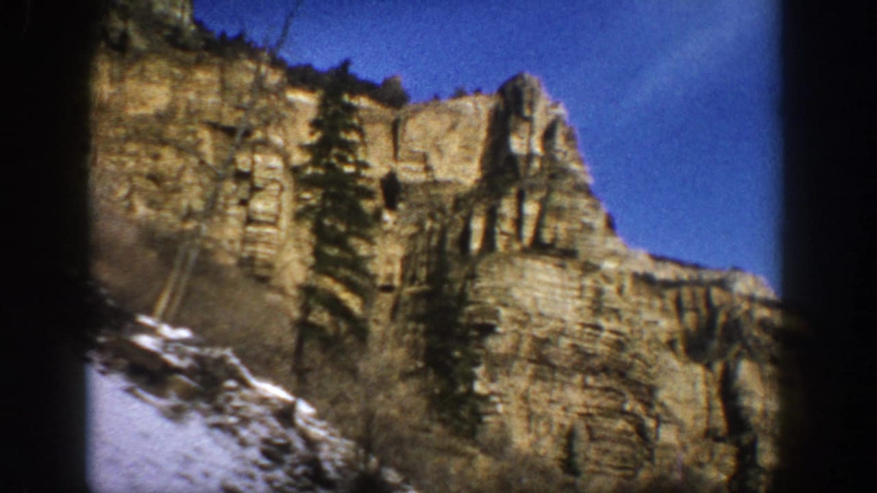 1961 Aspen-area 8mm frame showing a layered rocky cliff, sparse evergreens, blue sky, and snow patches at the lower slope.