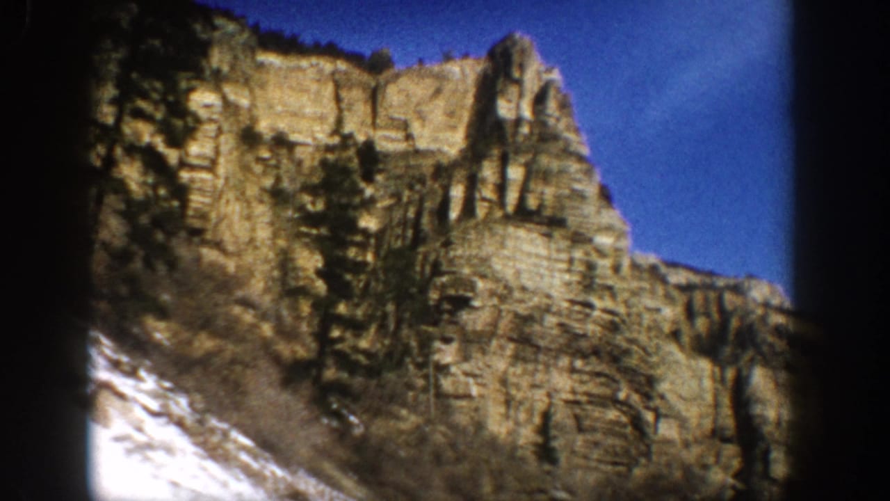 1961 Aspen-area 8mm view of a rugged cliff face with visible rock layers, scattered evergreens, snow at left, and clear sky.
