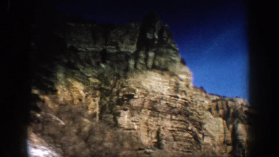 1961 Aspen-area 8mm frame with soft focus on a rocky mountain edge, brush, dark tree shapes, blue sky, and snow low at left.