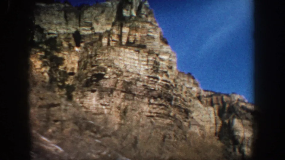 1961 Aspen-area 8mm frame showing the cliff face more tightly, with horizontal rock strata, trees, brush, and bright blue sky.