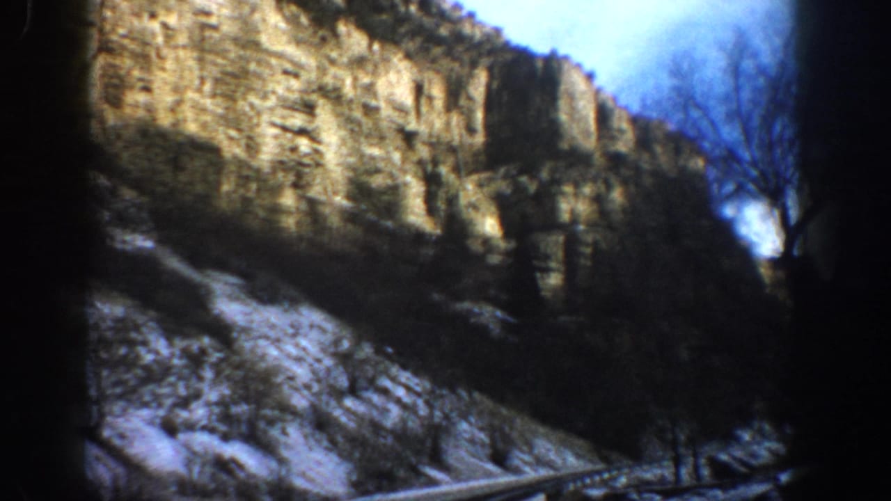 1961 Aspen, Colorado 8mm frame showing a snowy paved road beneath tall rocky cliffs and bare trees.