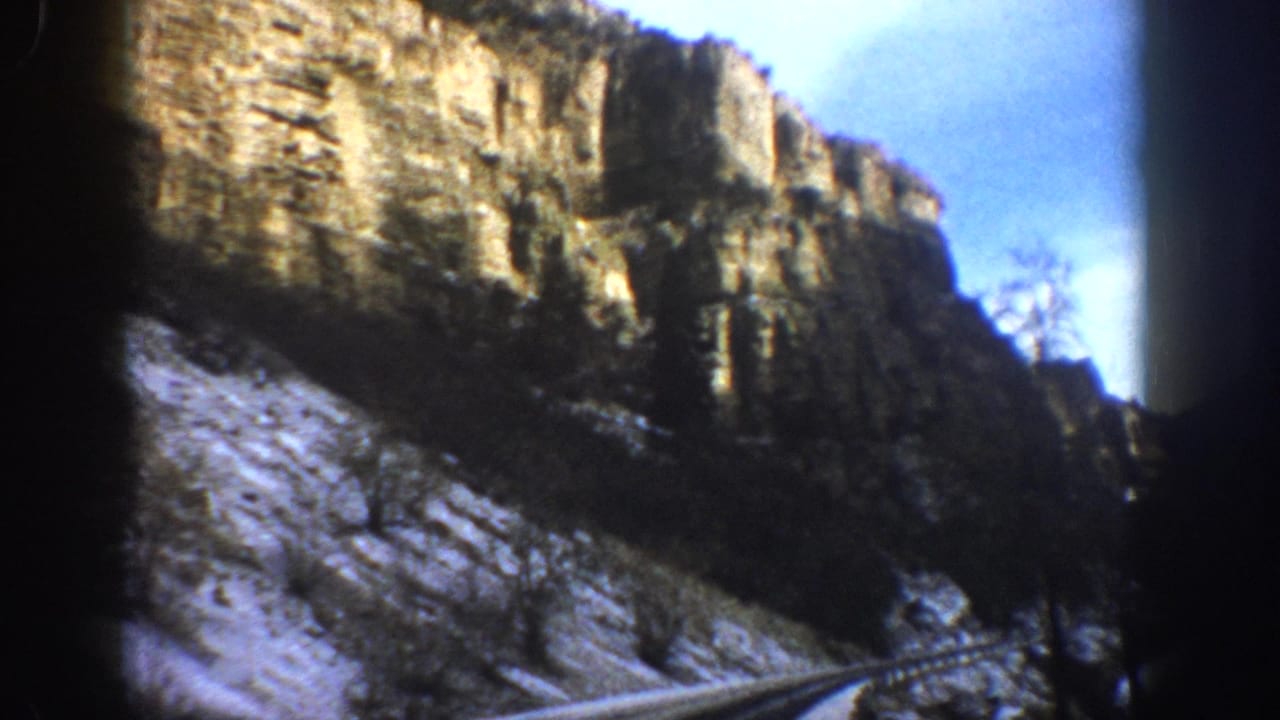 Curving snowy road bordered by cliffs, bare trees, and blue sky.