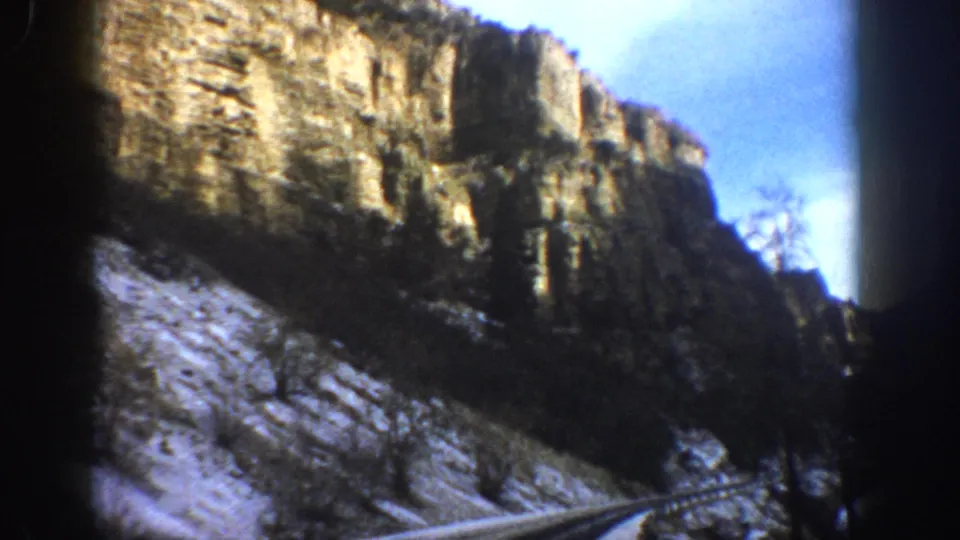1961 Aspen, Colorado travel footage frame with a paved road curving past snow-covered slopes and canyon cliffs.