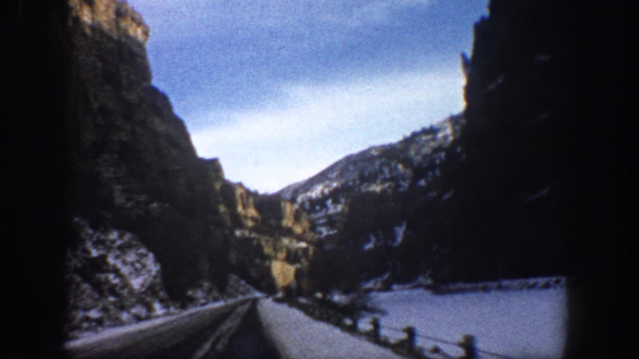 Roadside canyon view with snow, cliffs, guardrail, and distant hills.