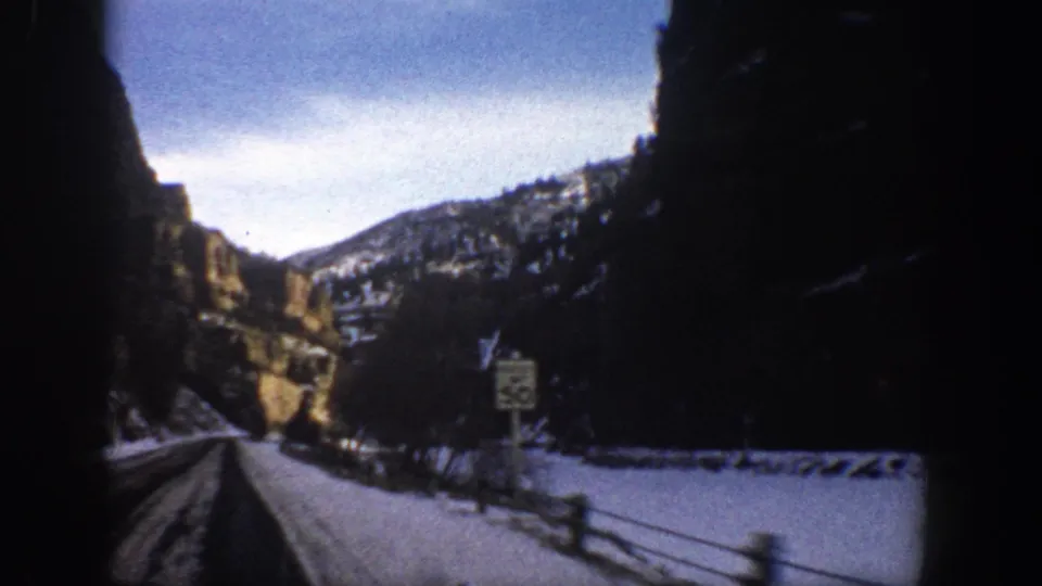 1961 Aspen, Colorado road-trip frame showing a snow-lined canyon road and a visible speed limit sign beside the shoulder.