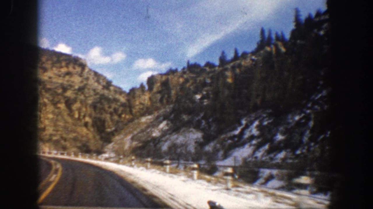 Curving road with snowbanks, guardrail, cliffs, and blue sky.