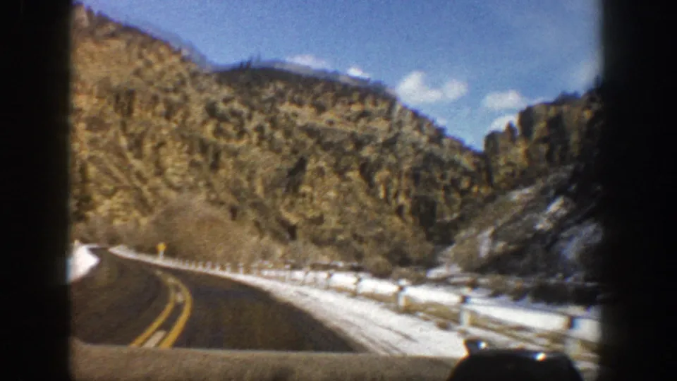 1961 Aspen-area mountain road footage with snowbanks, guardrails, rocky hillsides, and evergreen trees visible ahead.