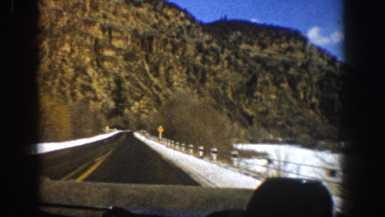Snowy highway curving toward rocky slopes with guardrail and road sign.