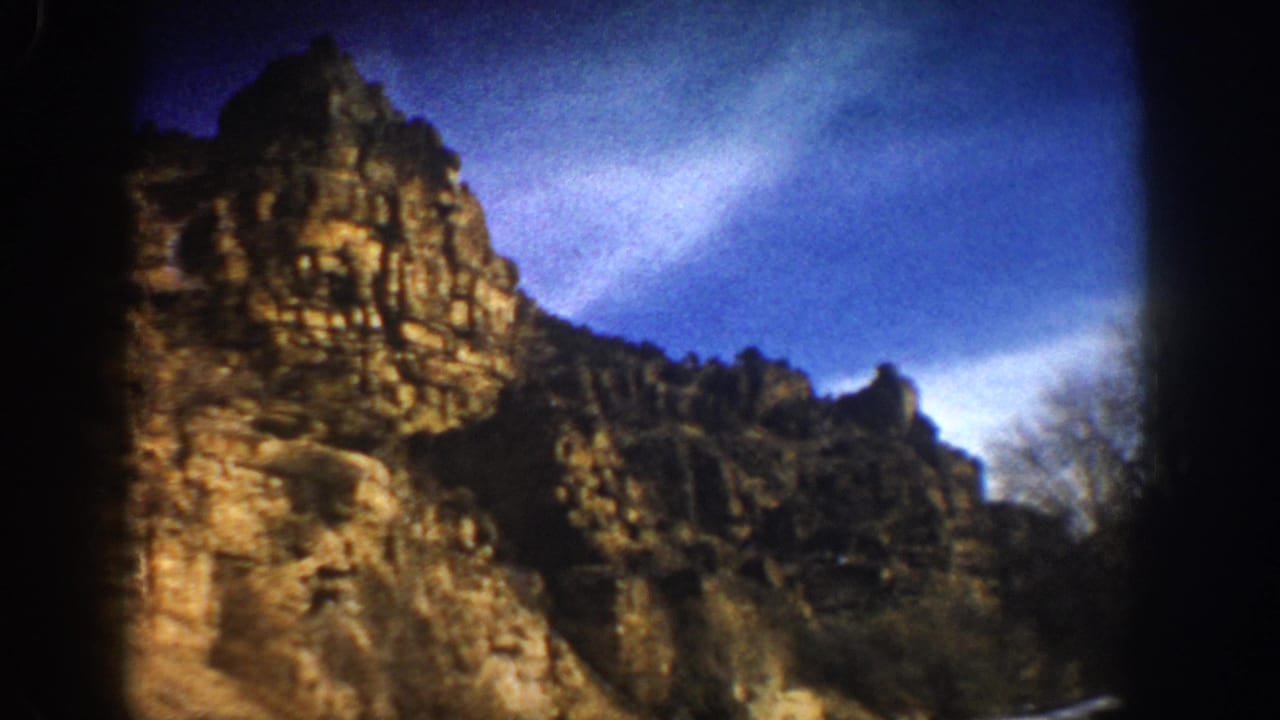 1961 Aspen-area 8mm frame showing a rugged cliff face beside a road under blue sky, viewed from a moving vehicle.