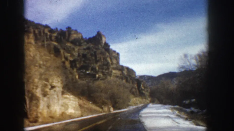 1961 Colorado mountain-road frame showing a wet two-lane road, snow patches, and rugged cliff faces ahead.