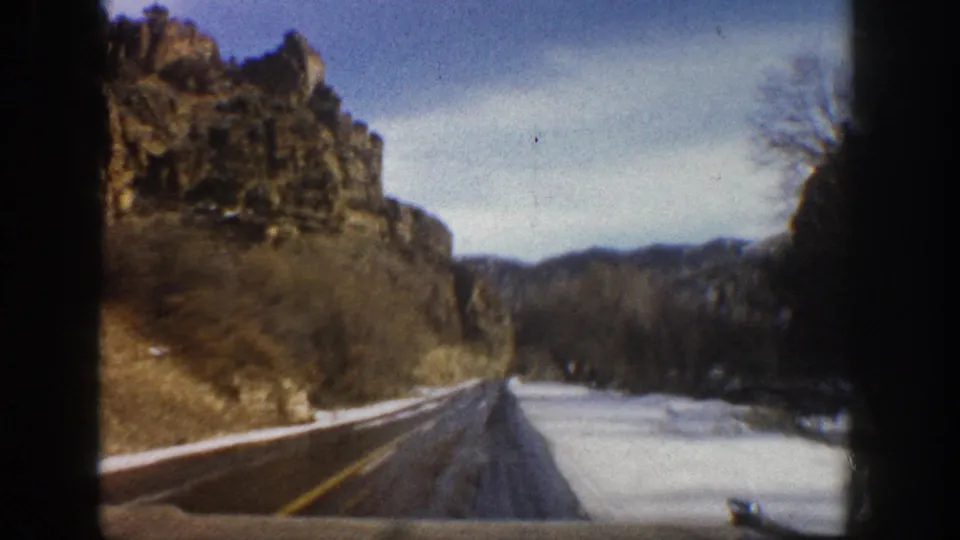Aspen-area 8mm frame showing a wet roadway through a snowy canyon with rocky cliffs and distant hills.