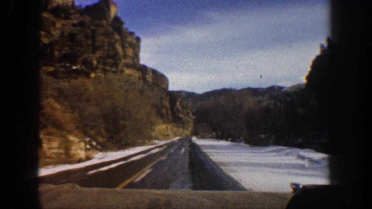 1961 Aspen-area 8mm frame from a moving vehicle, showing a mountain road, canyon walls, bare trees, and roadside snow.
