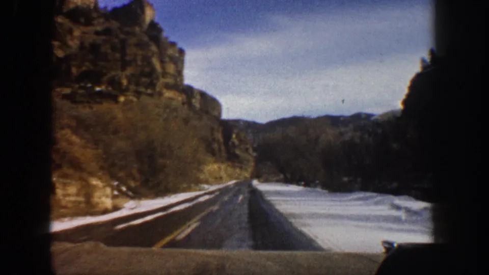 1961 Aspen-area 8mm frame from a moving vehicle, showing a mountain road, canyon walls, bare trees, and roadside snow.