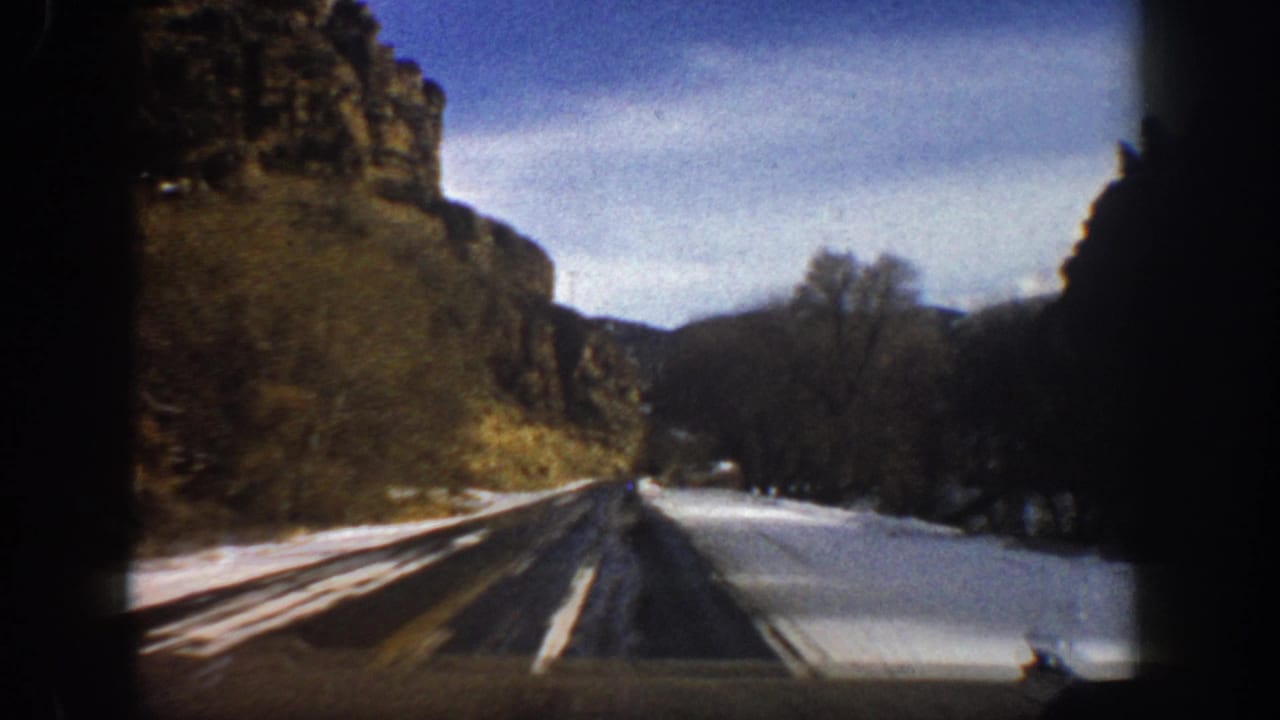 Moving-car 8mm view near Aspen, Colorado, with a winding road, snow patches, cliff faces, and winter-bare trees.
