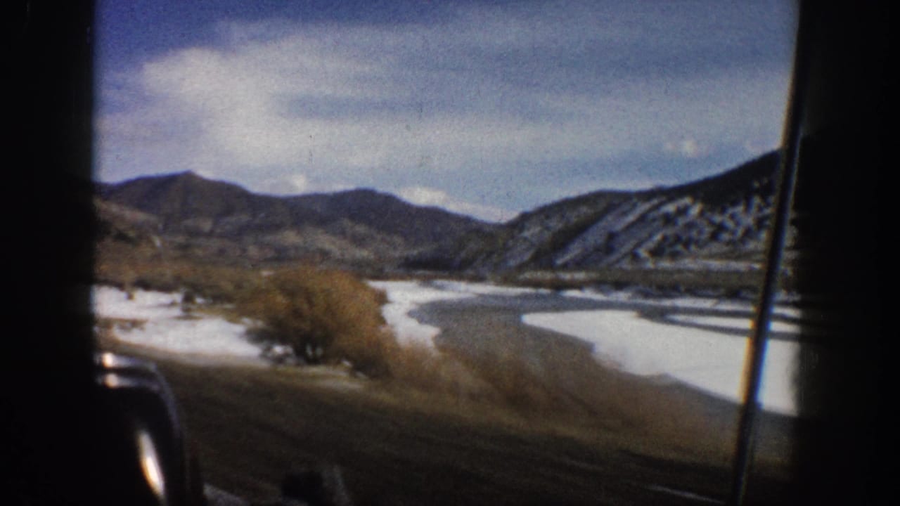 1961 Aspen, Colorado home-movie view from a moving vehicle, showing a river bend, snow-dusted ground, shrubs, and distant mountains.