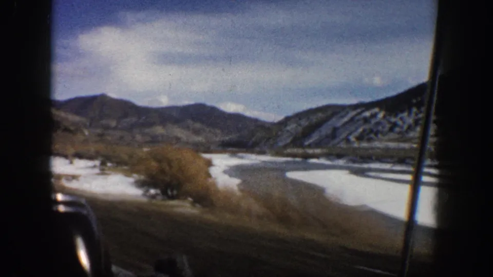1961 Aspen, Colorado home-movie view from a moving vehicle, showing a river bend, snow-dusted ground, shrubs, and distant mountains.