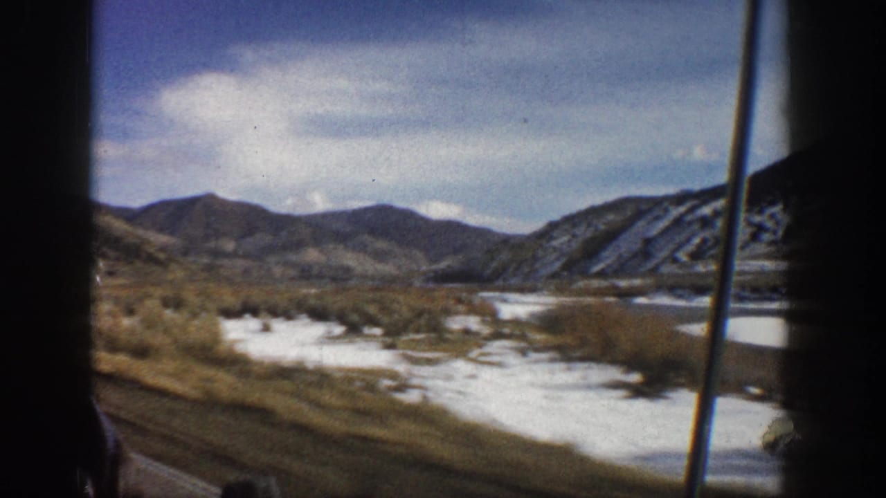 Moving-vehicle view near Aspen, Colorado in 1961, with dark window framing, snow along the valley floor, a river, and rugged hills beyond.