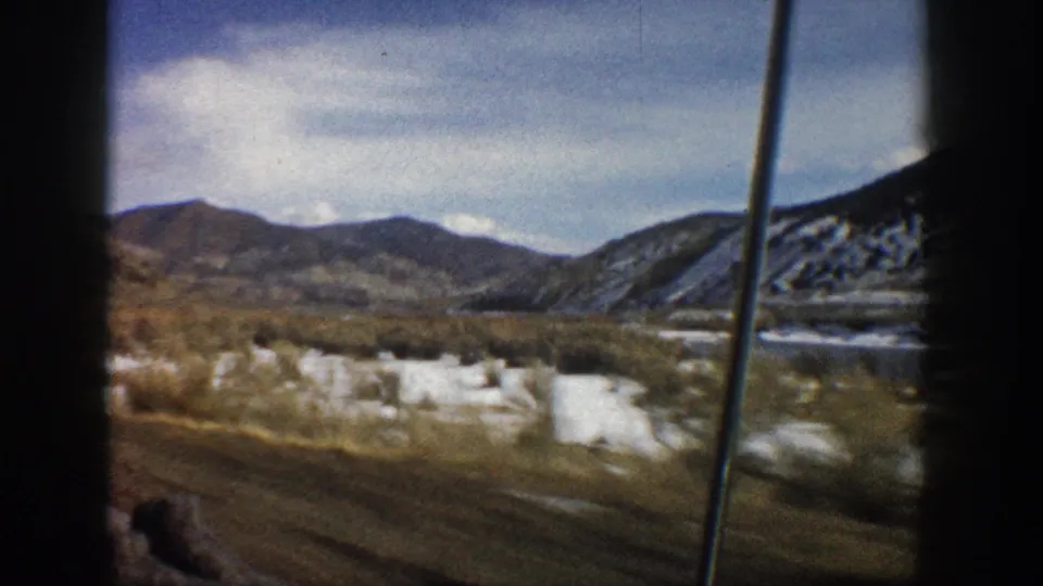 Aspen-area 1961 travel footage shows a blurred passenger-window view of snow patches, brush, a valley floor, and distant mountain ridges.